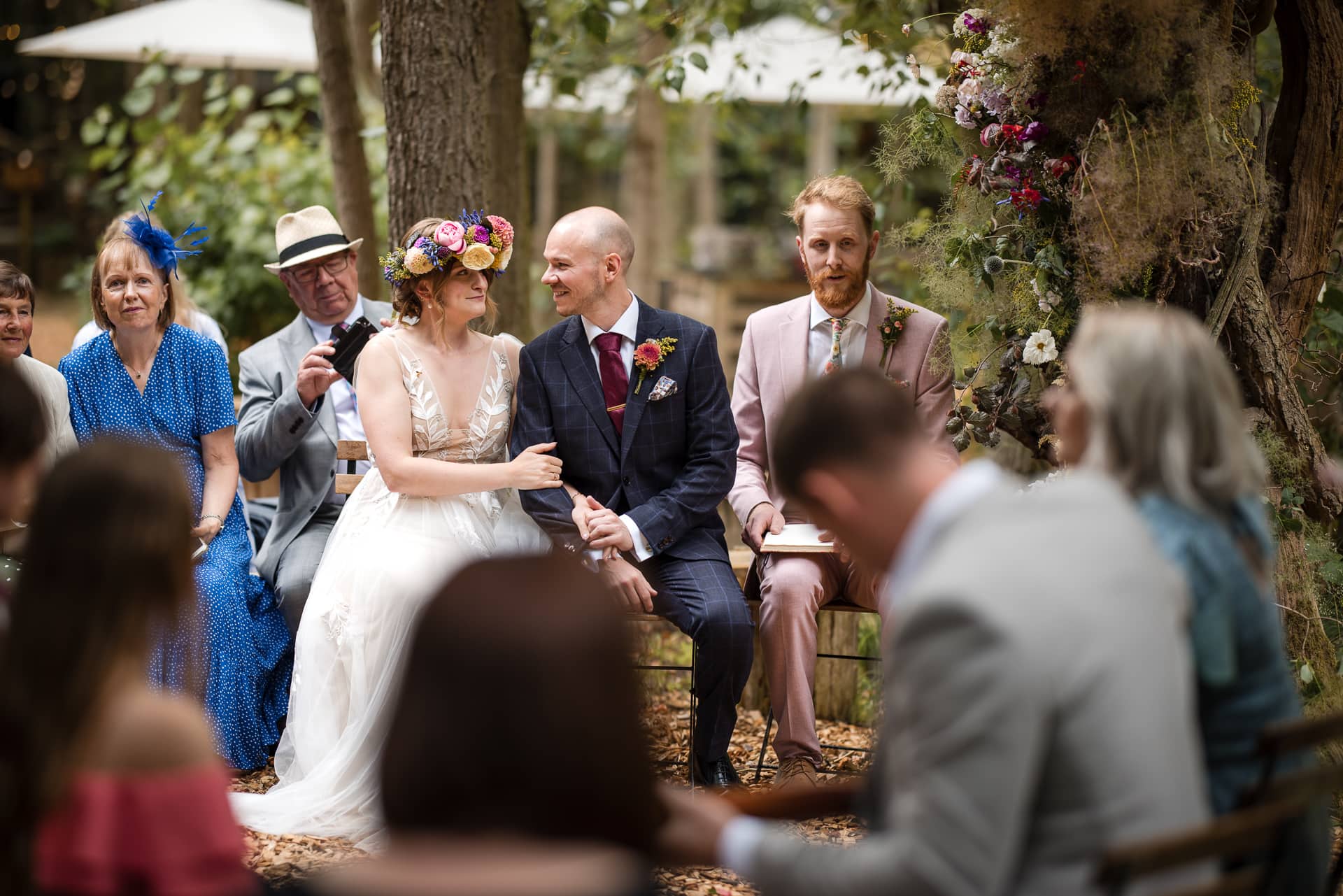 Bride and Groom sat looking at each other whilst friends sing a song at an outdoor woodland wedding.