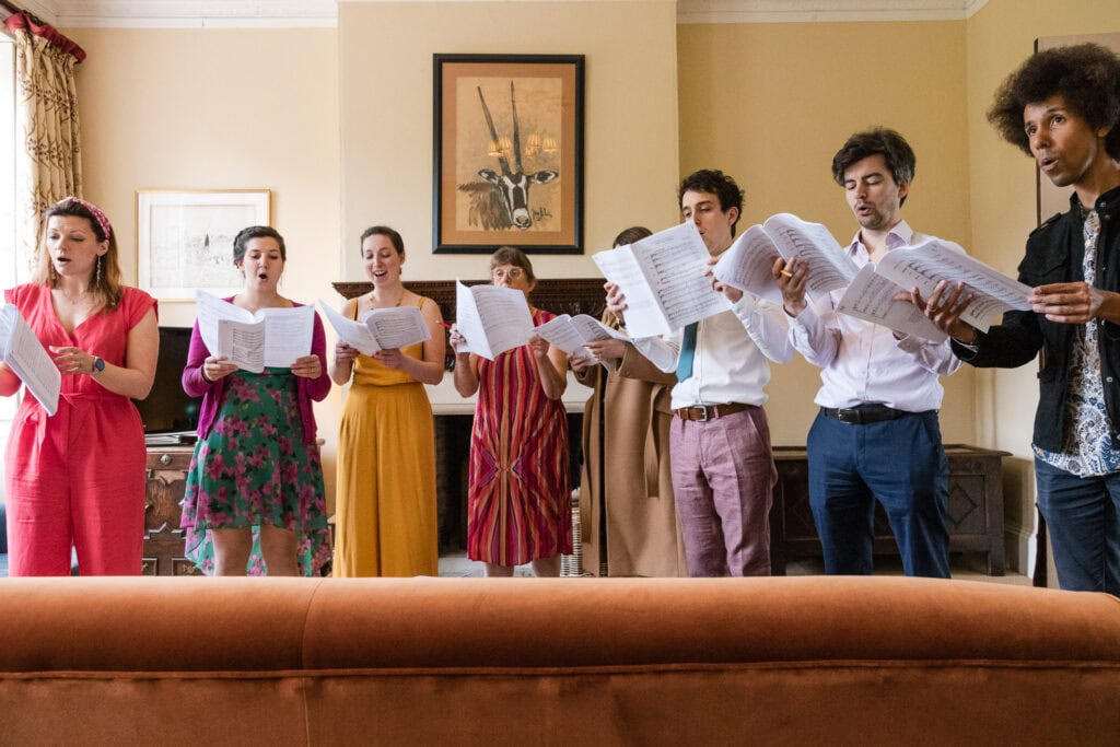 A group of eight people stand in a room, holding sheets of paper and singing. They are arranged in a line, facing forward, with a variety of expressions and outfits. The room is warmly lit, with framed art and a cushioned sofa in the foreground.