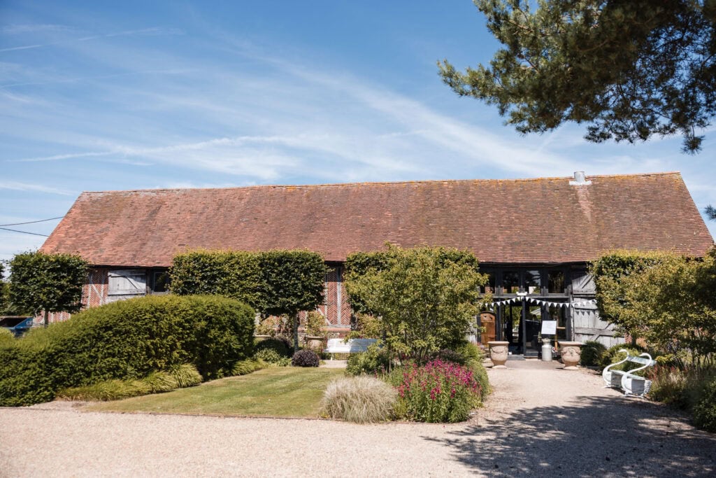 A rustic barn with a weathered red roof sits under a clear blue sky. The barn is surrounded by lush greenery, including trimmed bushes, flowering plants, and trees. A well-maintained gravel path leads to the barn entrance, flanked by stone pots and benches.