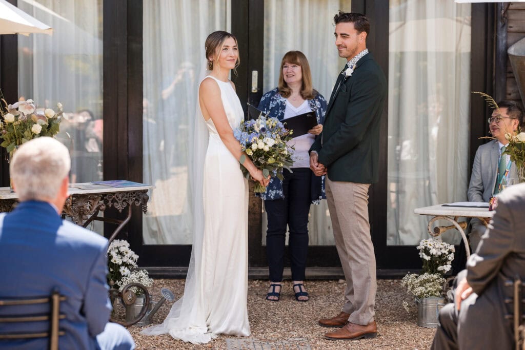A bride and groom stand facing each other, holding hands, during an outdoor wedding ceremony. The bride wears a white dress and holds a bouquet, while the groom is in a dark suit. An officiant stands behind them, and guests are seated on either side. Flowers decorate the scene.