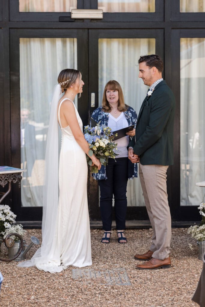 A couple stands facing each other during an outdoor wedding ceremony. The bride, wearing a white dress and veil, holds a bouquet. The groom, dressed in a suit, smiles at her. A woman officiant stands between them, holding a book. A glass door is in the background.