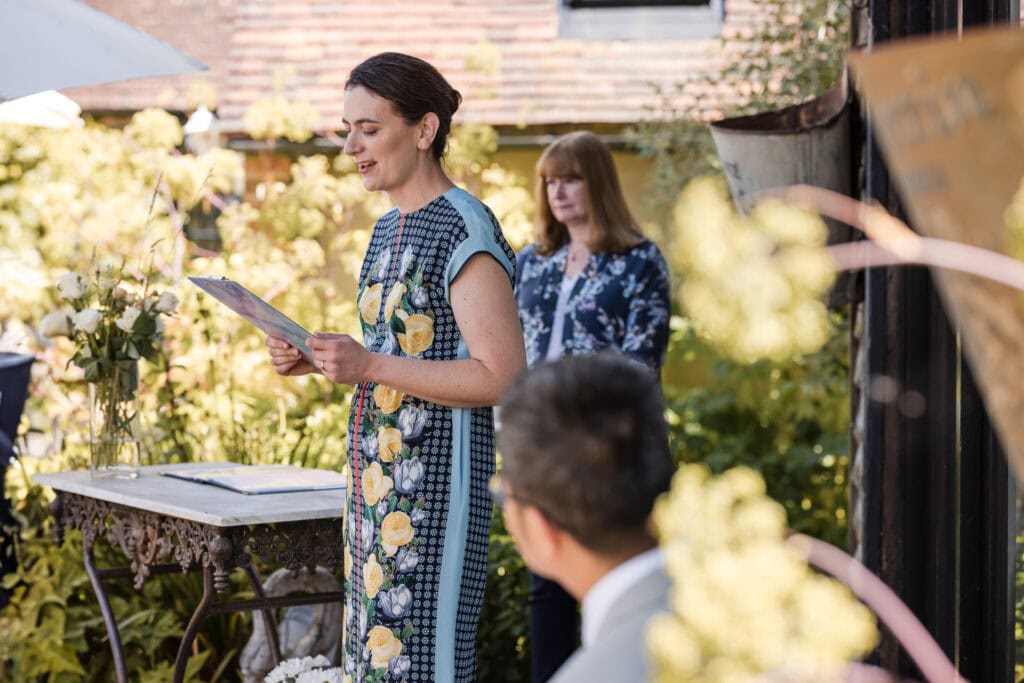 A woman in a floral dress is standing and speaking while holding a clipboard during an outdoor event. Another woman with long hair and a floral blouse stands in the background. Partially visible guests listen attentively, surrounded by greenery and rustic decor.