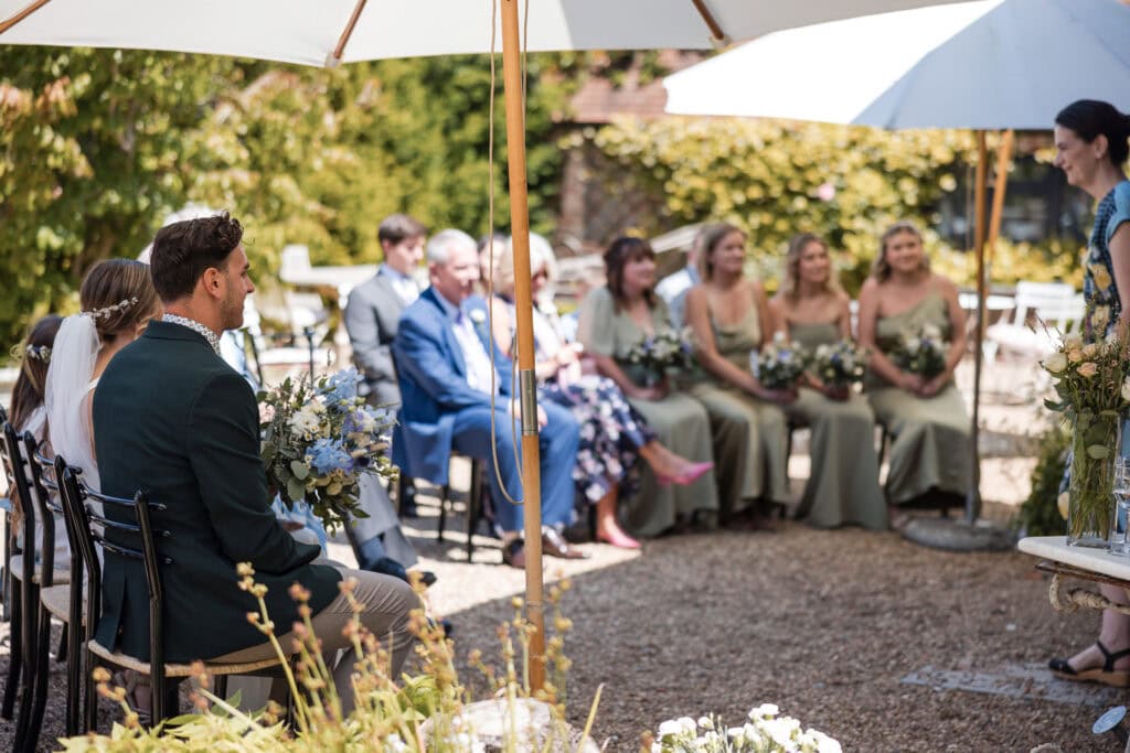 A wedding ceremony set outdoors. Guests are seated under white umbrellas, with a couple facing the officiant. Bridesmaids in green dresses hold floral bouquets, while other guests, including a man in a green suit, watch the proceedings.