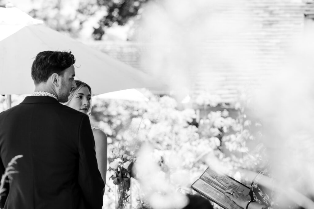 A black and white photo of a couple standing outdoors, facing partially away and listening intently to someone holding a book or folder. Trees and sunlight are seen in the background. The scene appears to be part of a formal or ceremonial event.