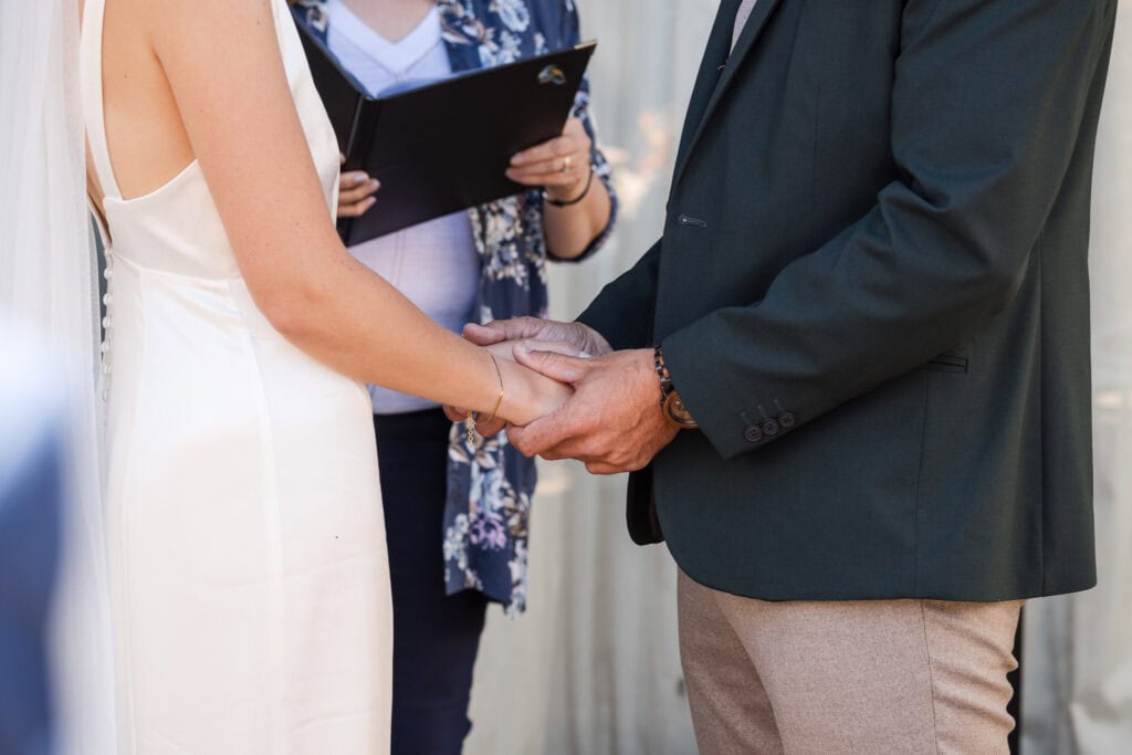 A couple holds hands during their wedding ceremony, with the officiant standing in the background holding documents. The bride is wearing a white sleeveless dress and veil, while the groom is in a green jacket and beige pants.