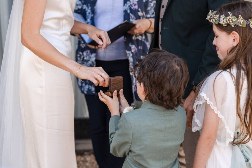 A bride in a white dress hands a wooden box to a young boy in a green suit. A girl with a flower crown watches beside him. Two adults stand in the background, one holding a book. The scene appears to be part of a wedding ceremony.