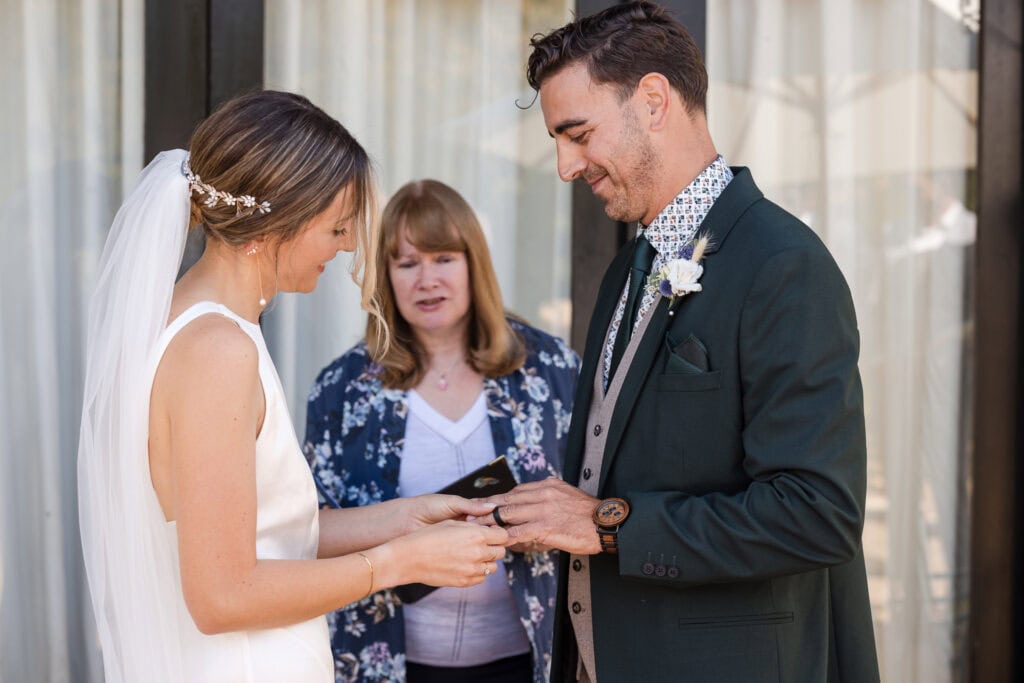 A bride and groom stand facing each other, holding hands during their wedding ceremony. The bride is placing a ring on the groom's finger. A woman stands behind them, possibly officiating the ceremony. They are outdoors with light-colored curtains in the background.