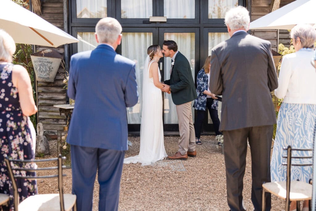 A bride and groom share a kiss in an outdoor wedding ceremony. The couple stands in front of a rustic building with glass doors. Guests, some seated and some standing, watch the moment from behind. White umbrellas provide shade over some of the chairs.