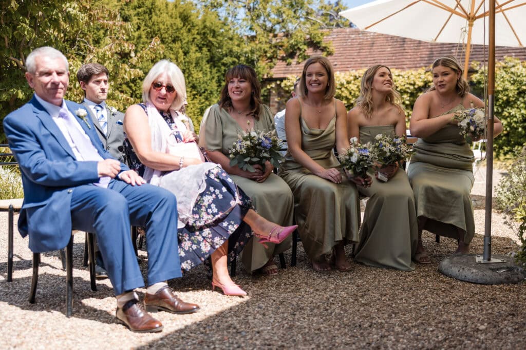 A group of people seated outdoors at a wedding ceremony. An older man in a blue suit and a woman in a floral dress with light pink heels sit on the left. Four bridesmaids in sage green dresses with bouquets are seated to the right, under a white umbrella.