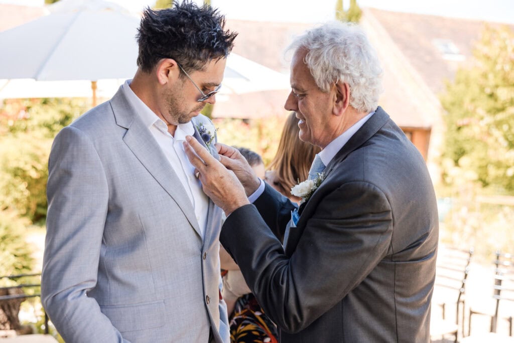 An older man helps a younger man adjust his boutonniere at an outdoor event. Both are wearing suits and sunglasses. The background shows a sunny day with a white umbrella and a building with a tiled roof.