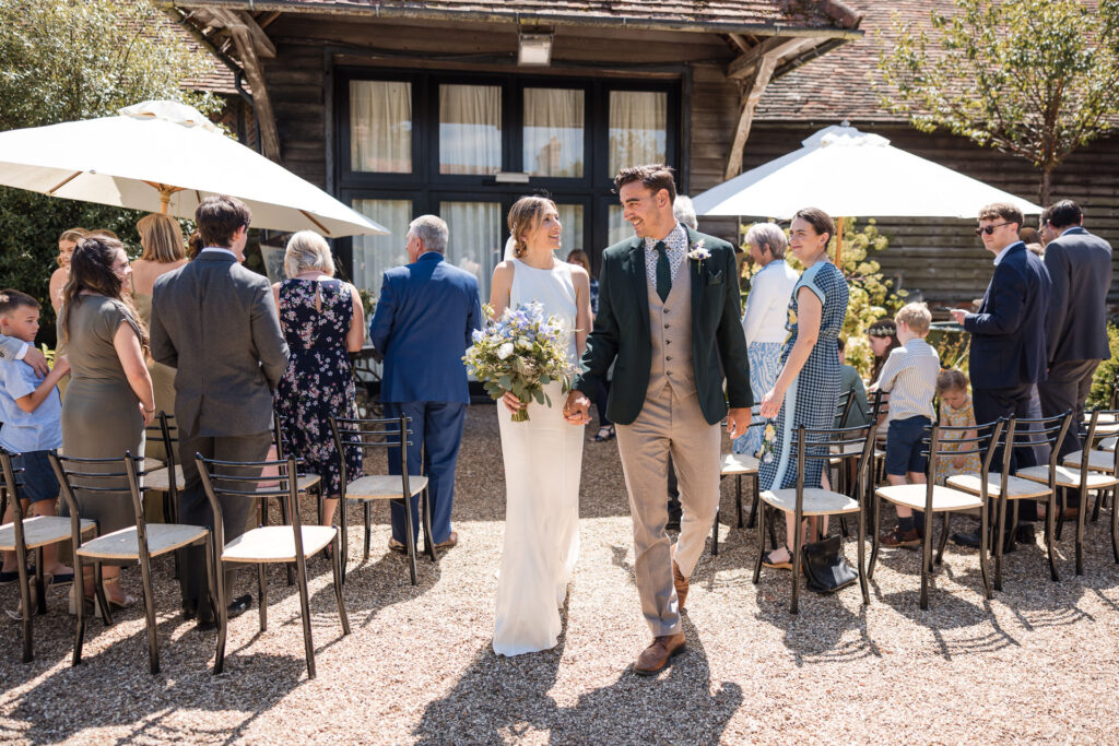 A bride and groom walking hand-in-hand down a gravel path at an outdoor wedding ceremony. The bride is in a white dress holding a bouquet, and the groom is in a suit. Behind them, guests are seated and standing, with white umbrellas providing shade.