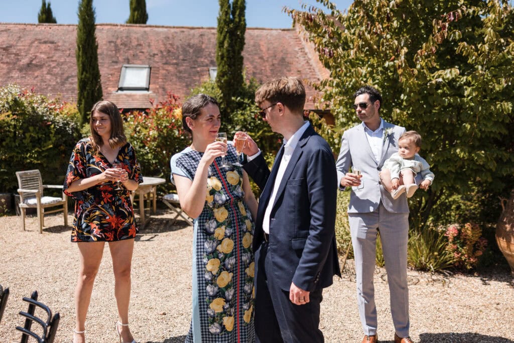 A group of five people are enjoying an outdoor gathering on a sunny day. A man in a suit and a woman in a floral dress are talking and holding drinks. Nearby, a man holding a baby and another woman in a patterned dress stand and chat, all surrounded by greenery.
