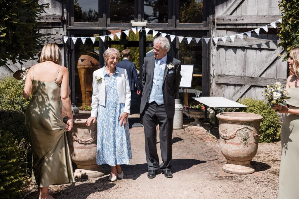 A group of people stands conversing outside a rustic building decorated with white bunting. An elderly couple, dressed in formal attire, is at the center. The woman wears a light blue dress and white jacket, and the man wears a suit with a blue waistcoat. Flower vases adorn the scene.