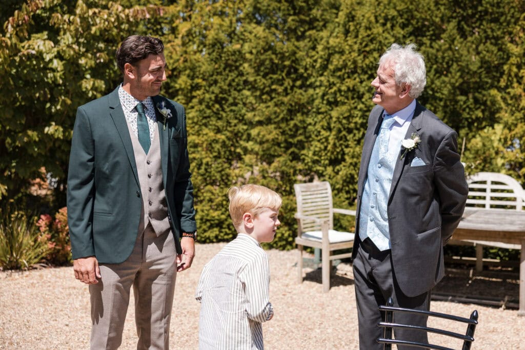 Three individuals, a younger man in a green jacket, an older man in a blue vest, and a young boy in a striped shirt, are standing outside having a conversation. They are in a sunlit garden area with trees, bushes, and garden furniture in the background.