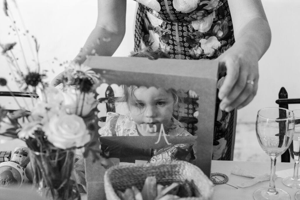 A black and white photo of a young girl sitting at a table with an open box in front of her. She is looking through the box, which has scalloped edges. An adult with a floral patterned dress stands behind her. The table has glasses, a basket of bread, and a vase of flowers.
