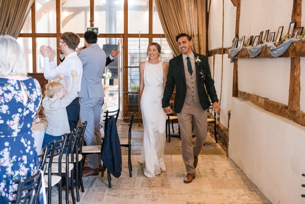 A newlywed couple, with the bride in a white sleeveless dress and the groom in a beige suit with a dark green jacket, walk hand-in-hand down an aisle, beaming smiles. Guests, including children, stand and clap in celebration inside a rustic, wooden venue.