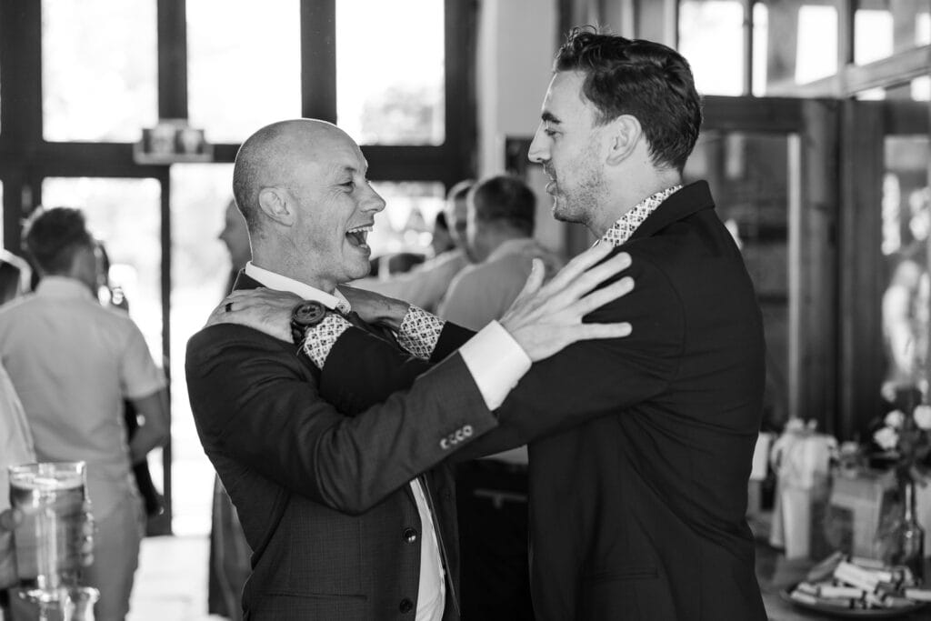 A black-and-white photo of two men in suits smiling and embracing each other in a lively indoor setting. The man on the left is bald, and the man on the right has short hair. Other people can be seen in the background, and the mood appears celebratory.
