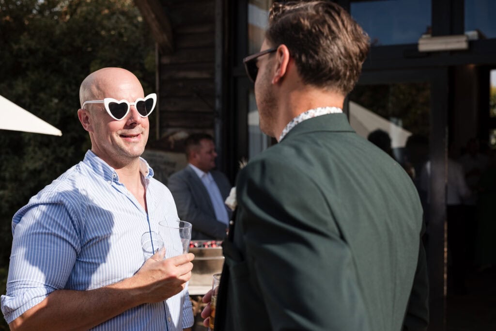 Two men are conversing at an outdoor event. The man on the left is wearing heart-shaped sunglasses, a light blue striped shirt, and holding a drink. The man on the right is wearing a dark jacket, sunglasses, and also holding a drink. They are smiling and engaged in conversation.