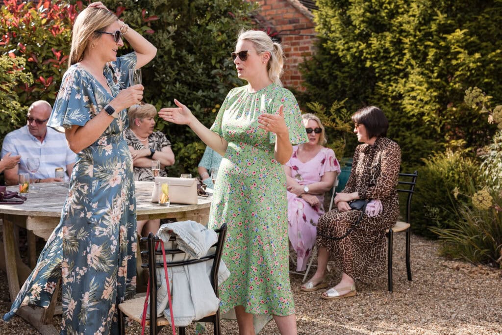 A group of people enjoying a garden party. Two women in floral dresses and sunglasses stand in the foreground having a conversation. Several people sit on chairs around a wooden table in the background, surrounded by greenery and plants.