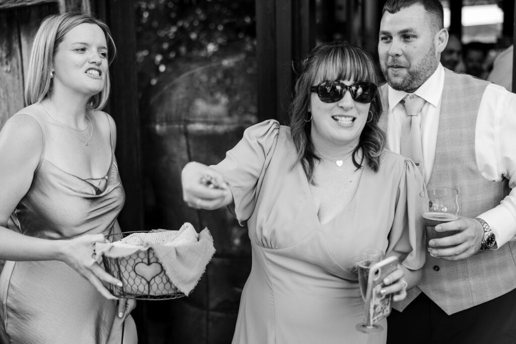 A black-and-white photo shows three people at what appears to be a festive gathering. One woman wearing sunglasses is enthusiastically gesturing while holding a drink. Another woman beside her is holding a basket, and a man in a tie and vest is holding a pint.