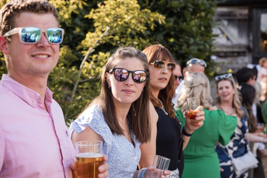 A group of people enjoying a sunny outdoor gathering. In the foreground, a man in a pink shirt and a woman in a light blue dress, both wearing sunglasses, hold drinks. Other attendees, also wearing sunglasses and holding beverages, are mingling in the background.