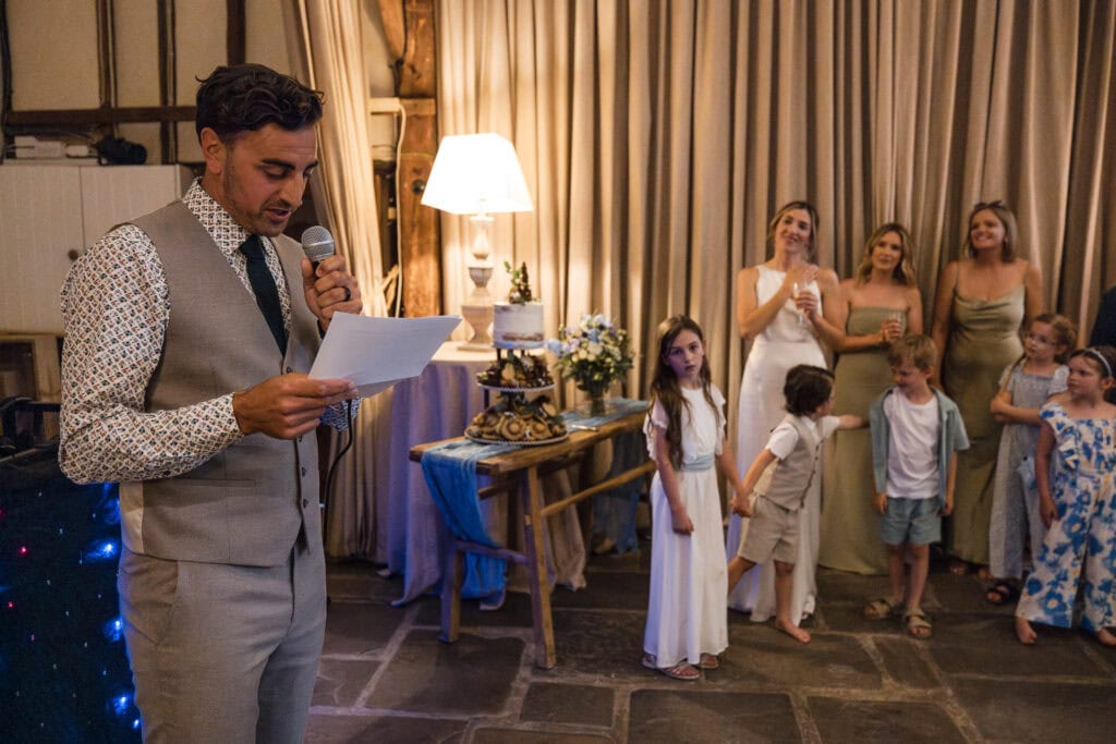 A man in a vest and dress shirt holds a microphone and reads from a piece of paper at an indoor event. Behind him, a group of women and children stand near a table with a lamp. Some women are smiling, and the children appear to be listening attentively.