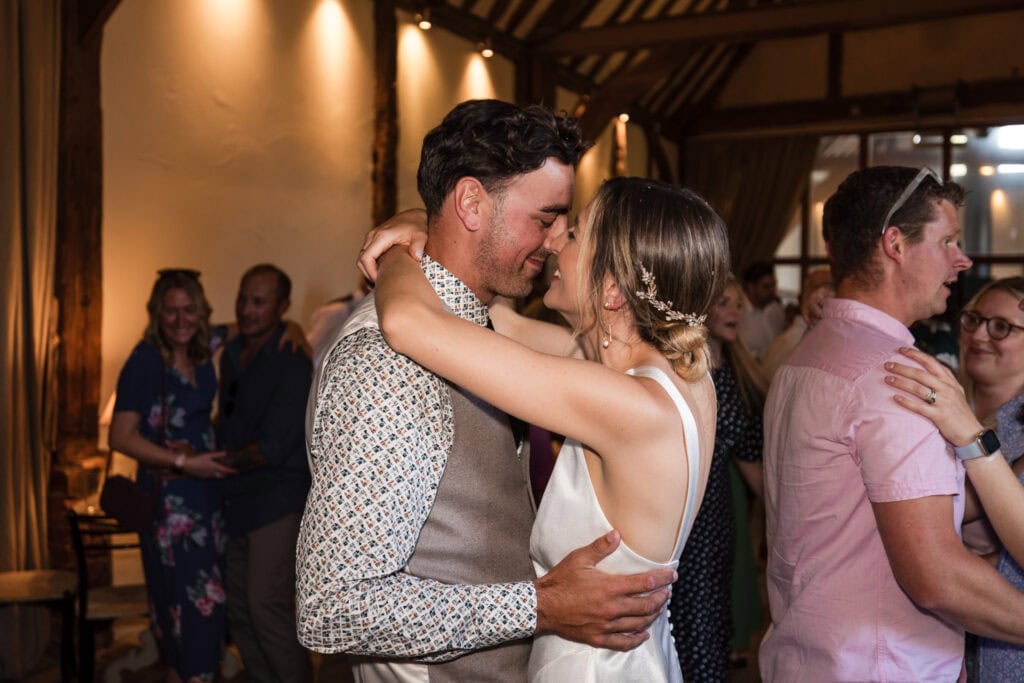 A couple shares a joyful dance at a warmly lit, rustic venue with exposed wooden beams. The man is in a patterned shirt and vest, while the woman wears a white dress. Other dancing couples and people mingling can be seen in the background.