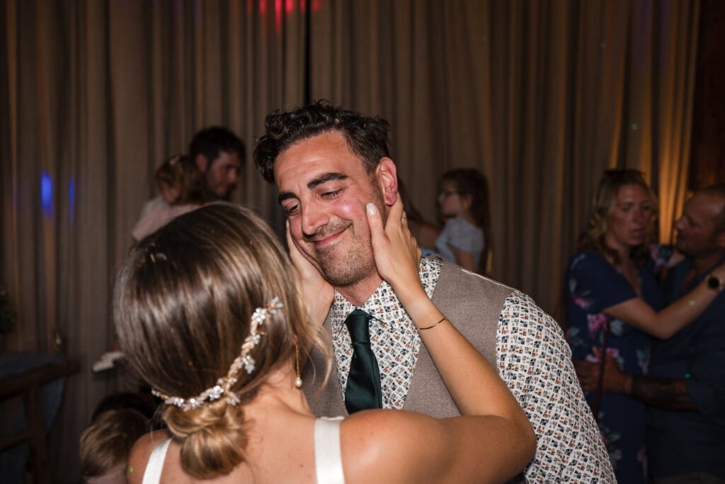 A couple is sharing a tender moment while dancing. The woman, back to the camera, wearing an elegant hair accessory, gently touches the smiling man's face. Other people and couples are in the background, indicating a festive atmosphere.