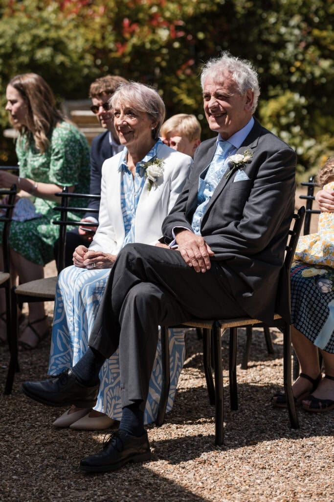 An elderly couple dressed in formal attire are seated outdoors at an event. The man is wearing a dark suit, and the woman is in a white jacket and blue dress. They are both smiling. Other attendees, including a woman in green, are visible in the background.