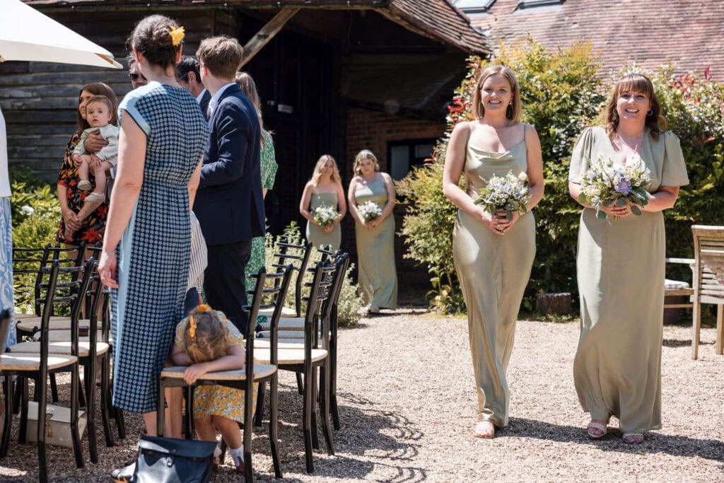 A group of bridesmaids wearing light green dresses walk down an outdoor aisle, holding flower bouquets. Guests, including a woman carrying a baby and a child leaning on a chair, observe while standing and sitting on black chairs under a sunny sky.