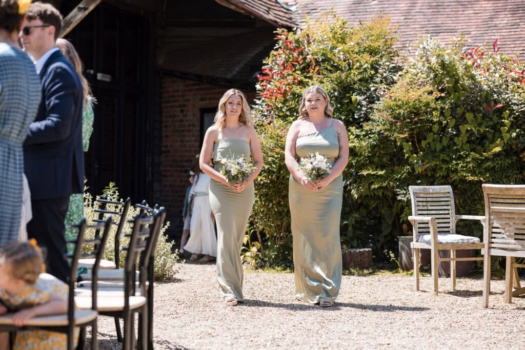 Two bridesmaids wearing light green dresses walk down an outdoor aisle holding bouquets at a sunny wedding ceremony. The aisle is lined with chairs, and guests are seated on either side. In the background, a building and lush greenery are visible.