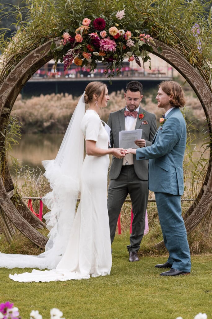 A bride in a white dress and a groom in a blue suit exchange vows outdoors, standing before a circular wooden arch adorned with flowers. An officiant in a gray suit stands between them. The scene is set by a river with a bridge in the background.
