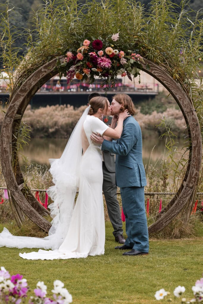 A bride and groom share a kiss at their outdoor wedding ceremony. They're standing in front of a large circular wooden arch adorned with colorful flowers. The bride wears a long white dress and veil, while the groom wears a blue suit.
