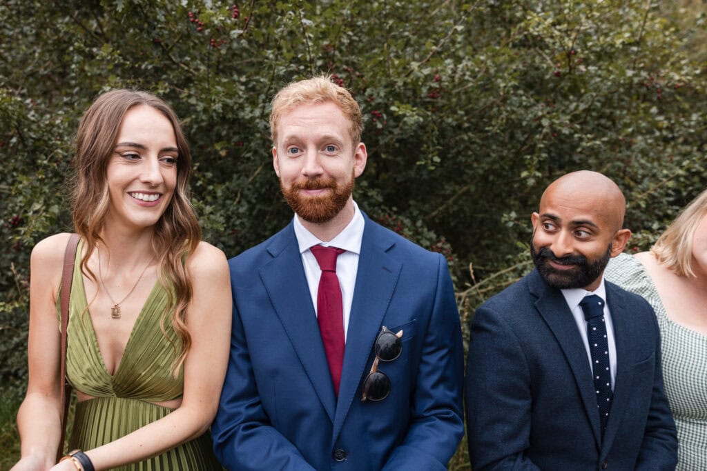 Three people are standing outdoors. A woman in a green dress smiles, a man in a blue suit with a red tie looks amused, and another man in a dark suit and polka dot tie smiles while looking at the man in the middle. Bushes are in the background.