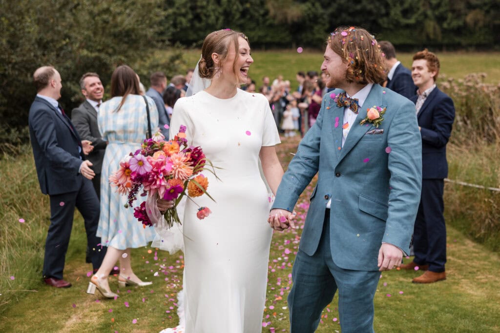 A couple joyfully walks hand in hand outdoors, surrounded by guests throwing confetti. The bride wears a white dress, holding a vibrant bouquet, and the groom is in a blue suit with a floral boutonniere. The scene is festive and lively.