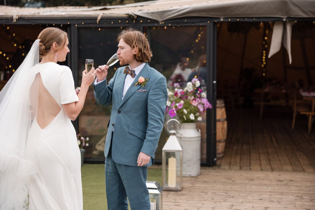 A bride and groom stand outside a venue with string lights, holding champagne glasses. The bride wears a white dress with an open back, and the groom is in a blue suit with a bow tie. Flowers and a decorative barrel are in the background.