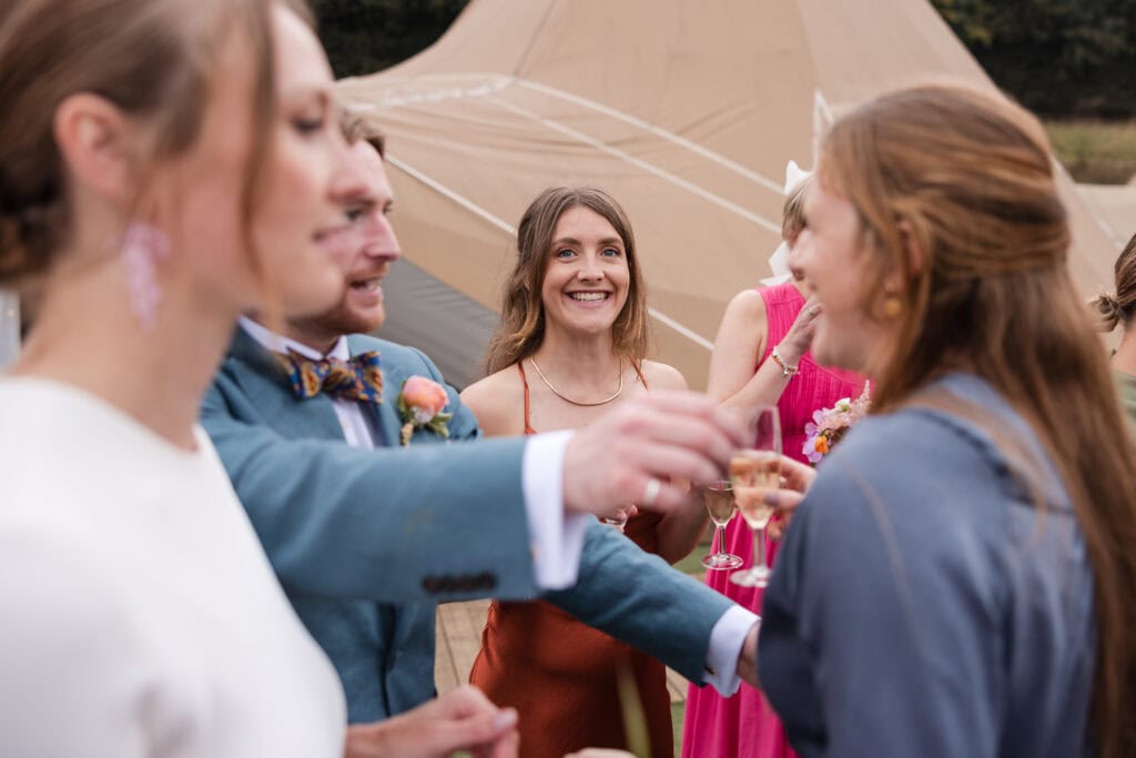 A group of people at an outdoor event, some holding glasses of champagne. The background features a large beige tent. One woman in an orange dress smiles in the center. The atmosphere seems lively and festive.