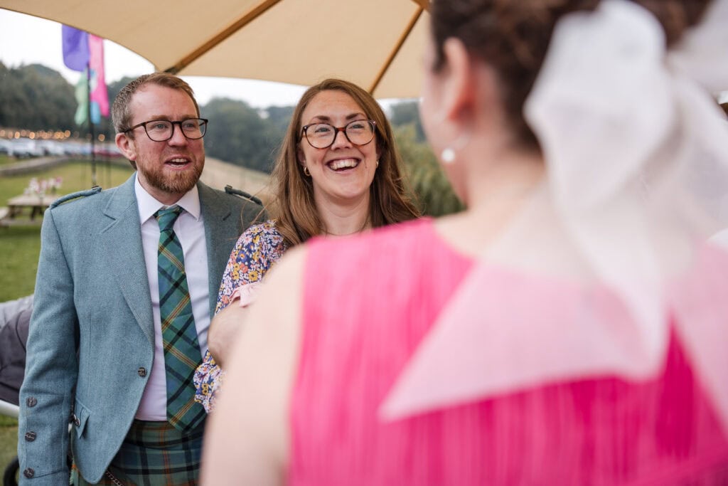A man and woman, both wearing glasses, smile at a person in a pink outfit with a white bow. The man wears a blue jacket and green plaid tie, and the woman wears a floral dress. They are outdoors under an umbrella.