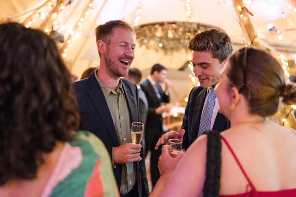 A group of people in formal attire are socializing and laughing at a festive event. One man holds a champagne glass. The background is decorated with lights and greenery, creating a warm and celebratory atmosphere.