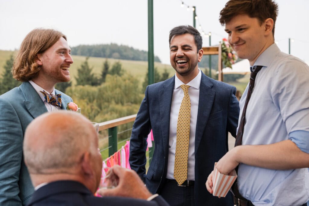 Four people are enjoying a conversation outdoors. Three are standing, dressed in suits, and one is seated with his back to the camera. They appear to be at a social event, with decorative bunting in the background and a scenic view of trees and hills.