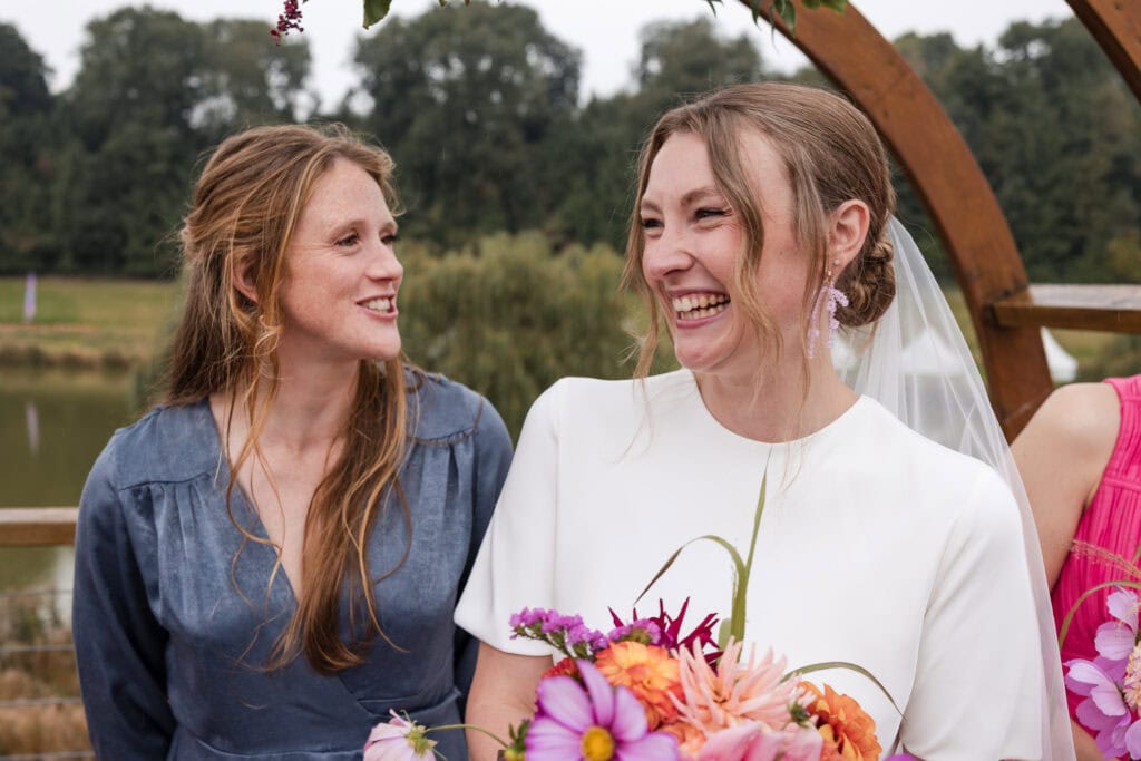 Two women are smiling and talking at an outdoor event. One is wearing a white dress and holding a colorful bouquet, while the other is in a blue outfit. They are near a wooden structure with trees and a pond in the background.
