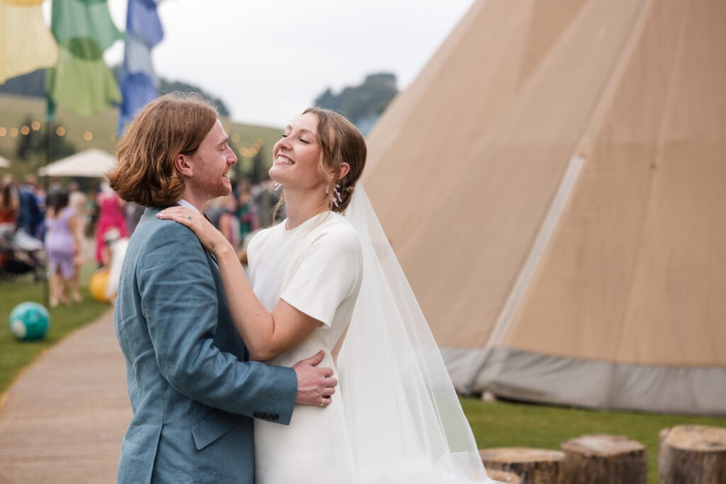 A joyful couple embraces at an outdoor wedding, standing in front of a large teepee. The bride wears a white dress with a veil, and the groom is in a blue suit. Colorful flags and blurred guests are visible in the background.