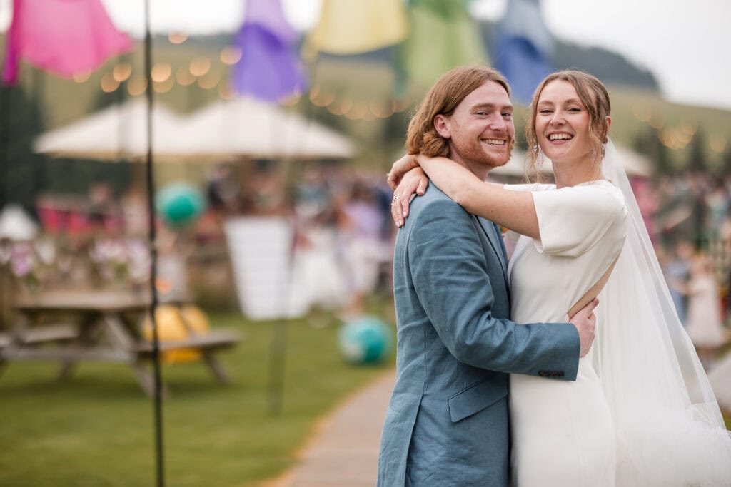 A smiling couple embraces outdoors at a wedding. The woman wears a white dress, and the man wears a light blue suit. Colorful flags and string lights decorate the background, with tables and guests visible in a festive, garden-like setting.