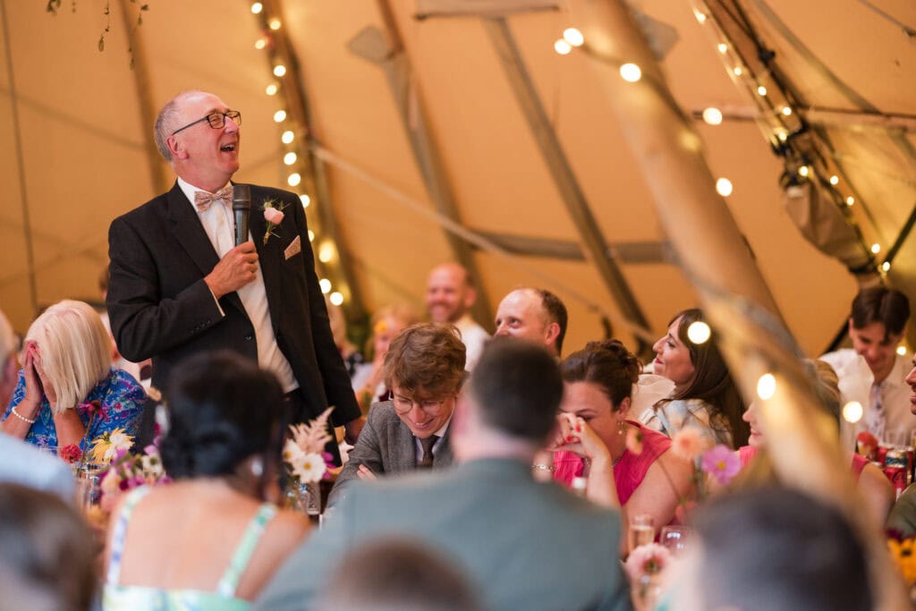 A man in a suit and bow tie stands and speaks into a microphone at a lively wedding reception inside a decorated tent. Guests sit around tables, smiling and enjoying the moment. Strings of lights add a festive atmosphere.