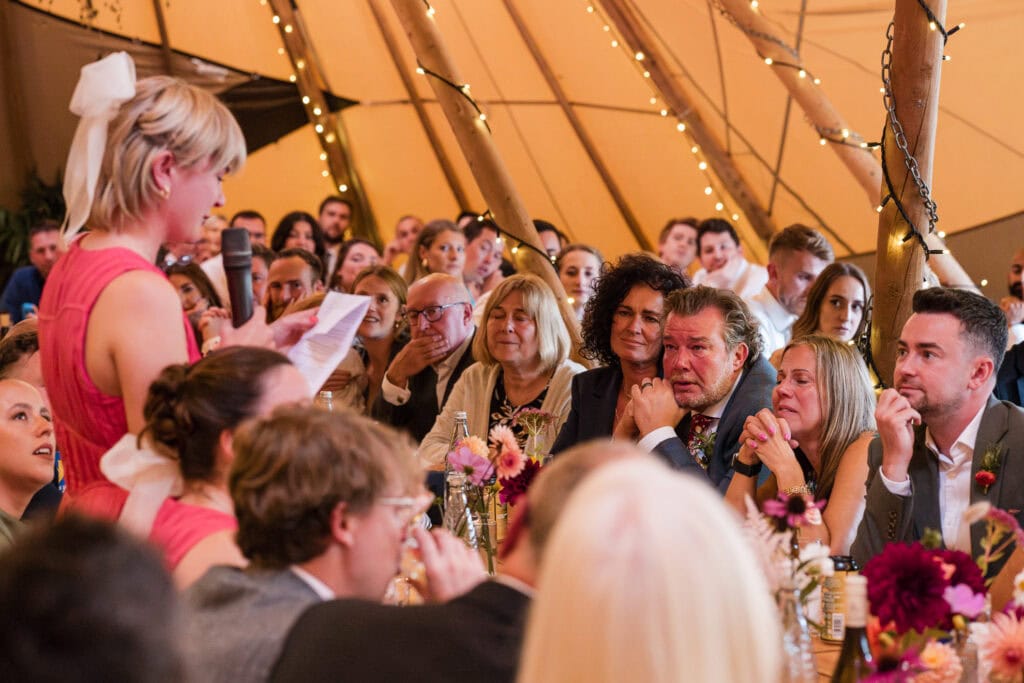 A woman in a pink dress and white bow speaks into a microphone at a crowded indoor event. Attendees sit around tables adorned with flowers, listening intently. The venue has a warm, festive atmosphere with string lights hanging from the ceiling.