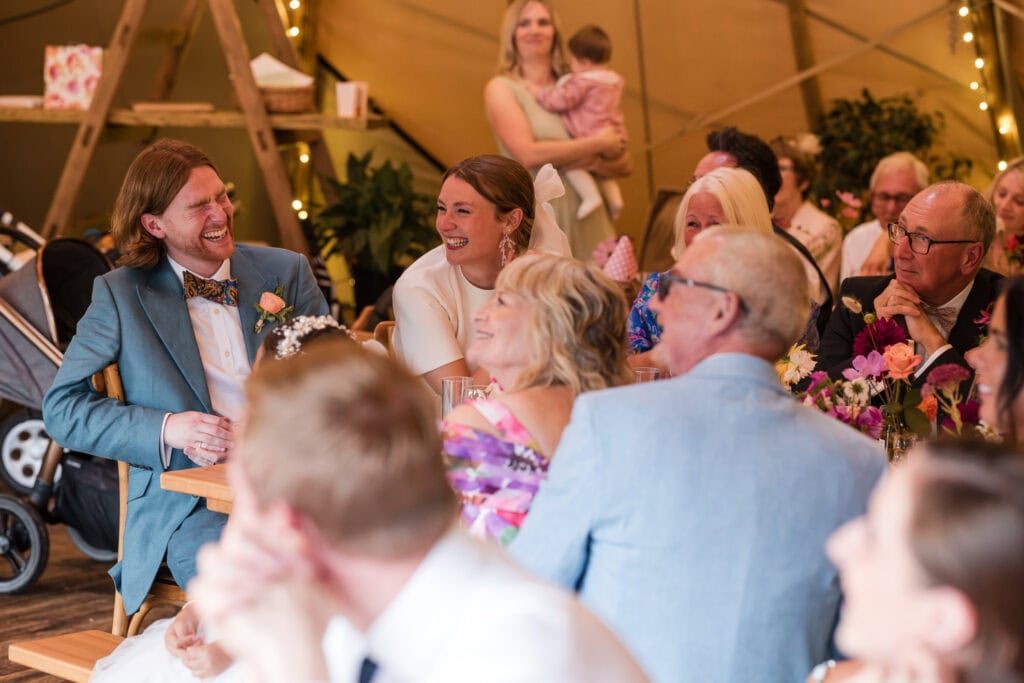 A joyful wedding reception scene with a smiling couple seated among their guests. The groom is in a light blue suit and the bride in white, surrounded by warmly dressed people in a rustic setting with a baby in the background.