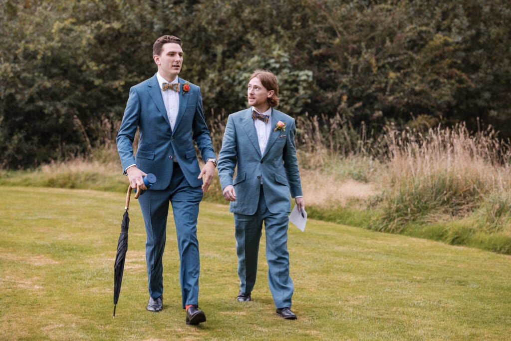 Two men in matching blue suits and bow ties walk on grass. One holds an umbrella, and they appear to be engaged in conversation. Tall grass and trees are in the background, suggesting a park or garden setting.
