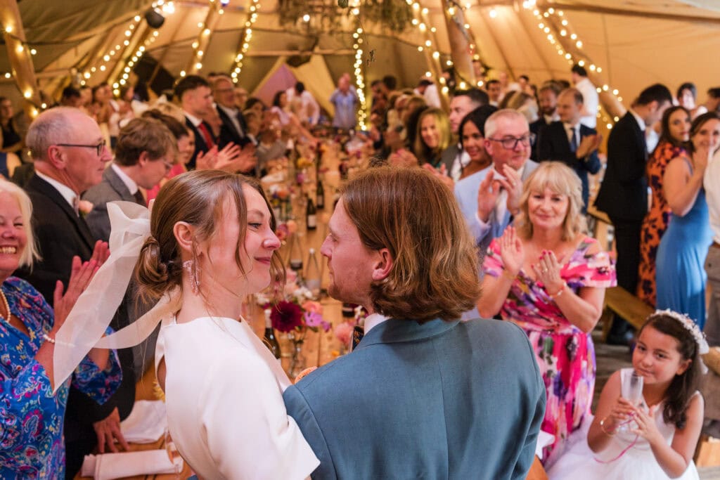A newlywed couple shares a joyful moment as they walk through a crowd of smiling, applauding guests inside a decorated tent. Strings of lights and floral arrangements add a festive touch to the celebration.