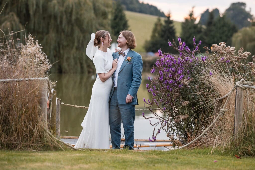 A couple in wedding attire stands on a wooden dock by a lake. They are smiling at each other, surrounded by purple flowers and tall grass. The background features a gentle hillside and trees.