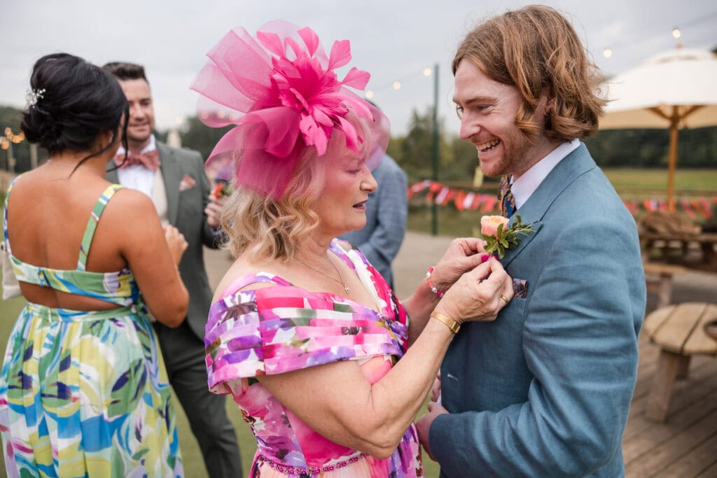 A woman in a colorful dress and pink hat pins a boutonniere on a man in a blue suit. They are outdoors, with two people in the background wearing summer attire at what appears to be a festive event.